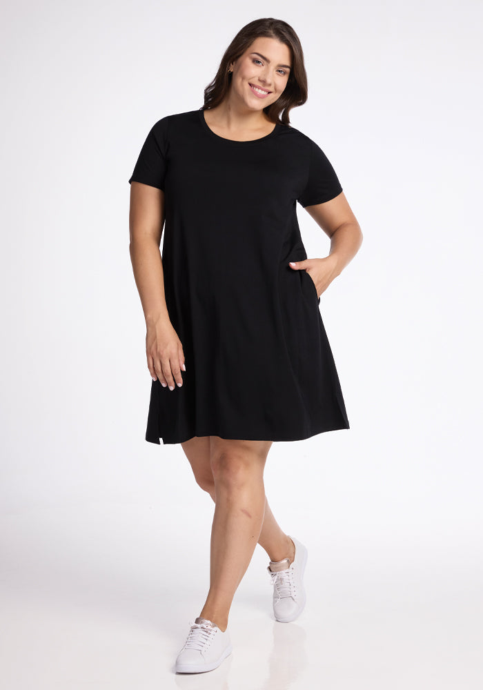A person smiles, standing against a plain white background, wearing the Georgie Dress - Black by Woolx. This A-line silhouette dress is crafted from merino wool and features short sleeves and pockets. Paired with white sneakers, they have one hand in their dress pocket. 