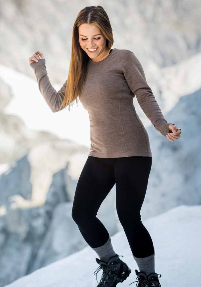 A woman with long brown hair, dressed in a brown long-sleeve shirt, heavyweight leggings, gray socks, and Woolx Stella Tall - Black boots, smiles while standing on snow with a blurred mountain backdrop.