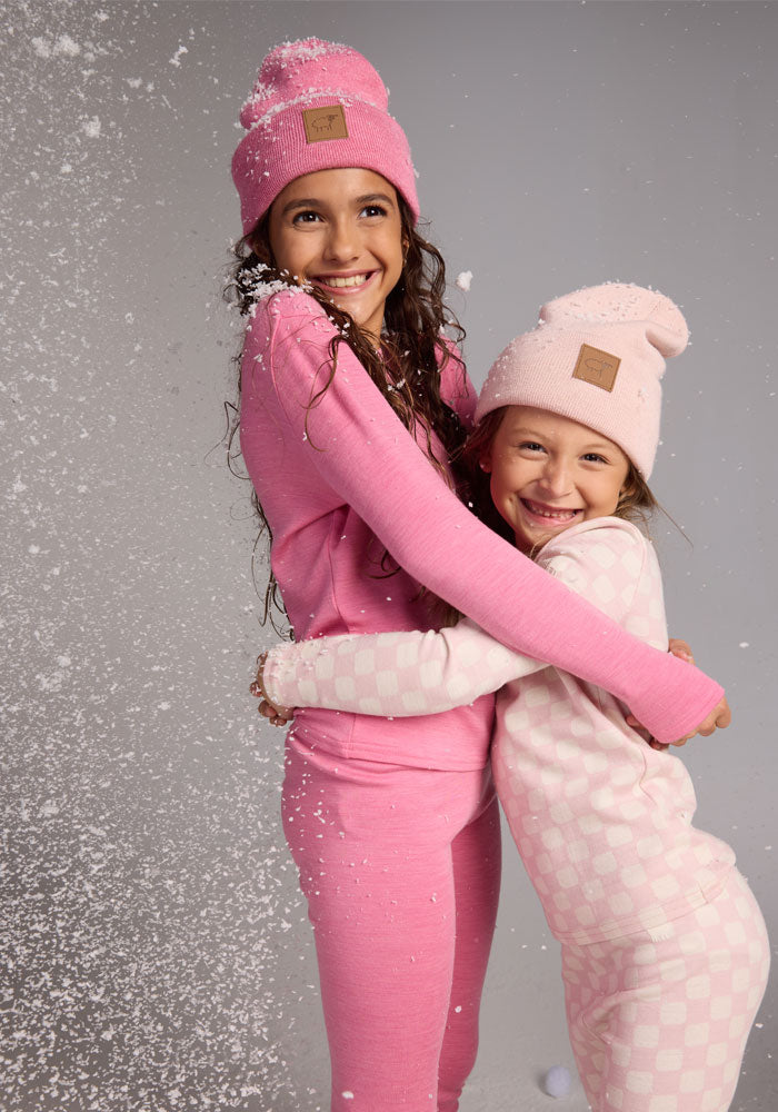 Two happy girls in pink Kids Explorer Base Layer Bottoms - Pinky Promise by Woolx hug as faux snow falls. Both with long hair, they smile brightly in matching pajamas and beanies against a plain gray backdrop.