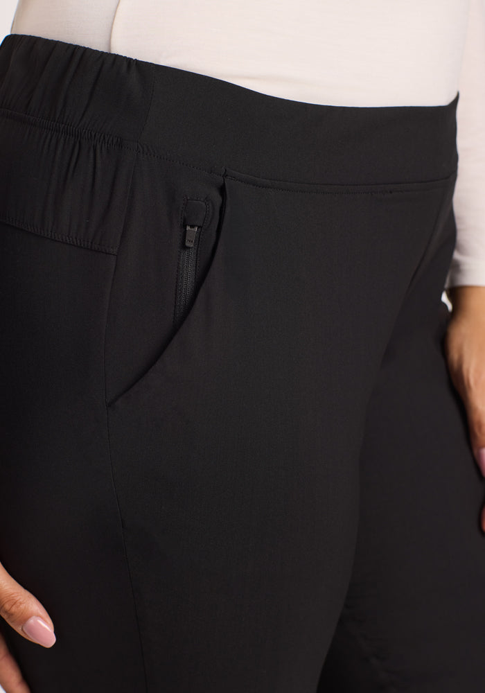Close-up of a person wearing Woolx Nicole Pants - Black, featuring breathable fabric, an elastic waistband, and a zippered side pocket. Their hand rests near the pocket, paired with a long-sleeve white top.