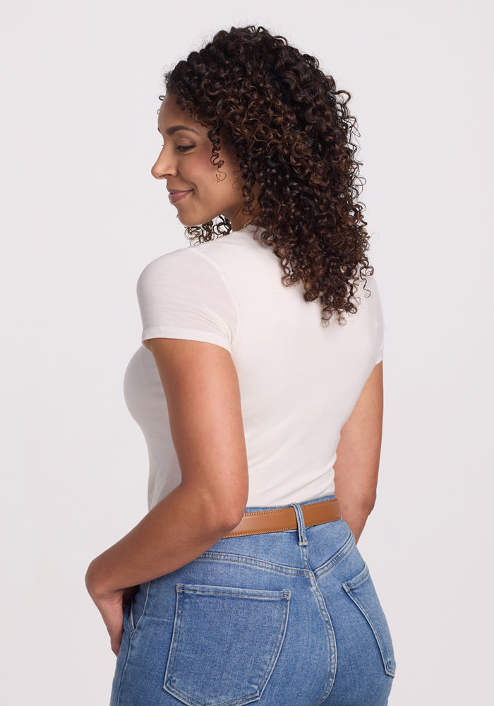 A woman with curly hair smiles over her shoulder, wearing the Woolx Gracie Bodysuit in Vanilla—a fitted, moisture-wicking Merino wool piece—paired with high-waisted blue jeans against a plain light background.