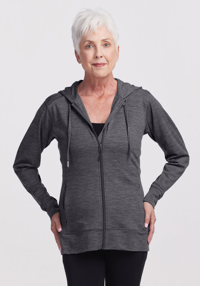 Older woman with short white hair wearing the Woolx Ryann Hoodie in Cinder Melange and black pants, standing hands on hips against a plain light background. 