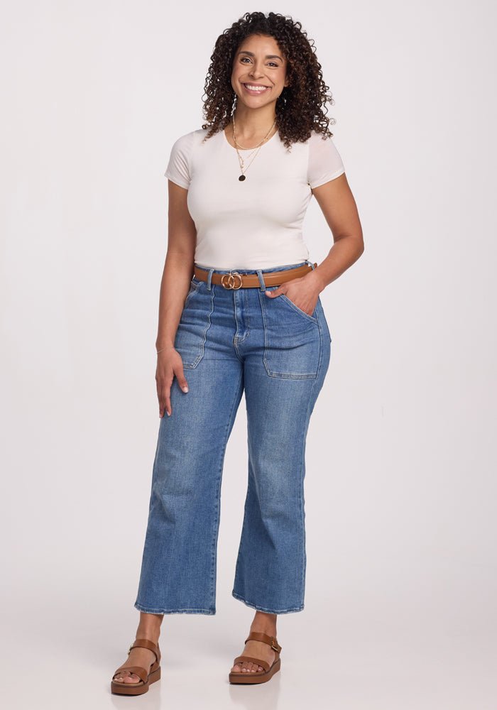 A woman with curly hair wears the Woolx Gracie Bodysuit in Vanilla under her white fitted t-shirt, blue high-waisted jeans, brown belt, and sandals, smiling against a plain white background.