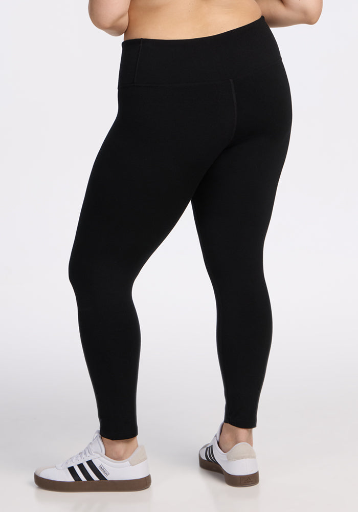 A person wearing black Woolx Angela Leggings and white Adidas sneakers stands against a plain white background, displaying the back and side views of the leggings.