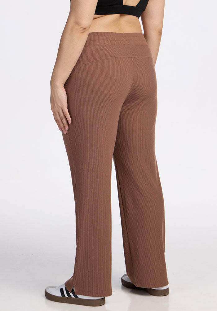 A person is seen from behind wearing Woolx's Luca Ribbed Pants in Sienna Kiss, paired with a black crop top and black-and-white slide sandals, standing against a plain white background.
