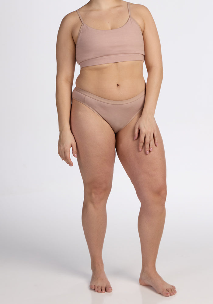 A woman stands barefoot against a plain white background, wearing the Woolx Luna Bralette - Morning Latte and matching underwear. Her right arm is by her side, her left hand on her thigh, and her face is not visible.