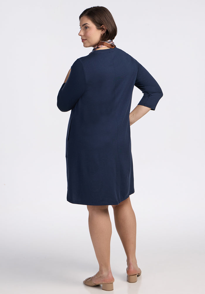 A woman faces away, wearing the Woolx Rissa A Line Dress in Deep Navy—knee-length Merino wool with three-quarter sleeves—styled with tan flats and a patterned scarf, set against a plain white background.