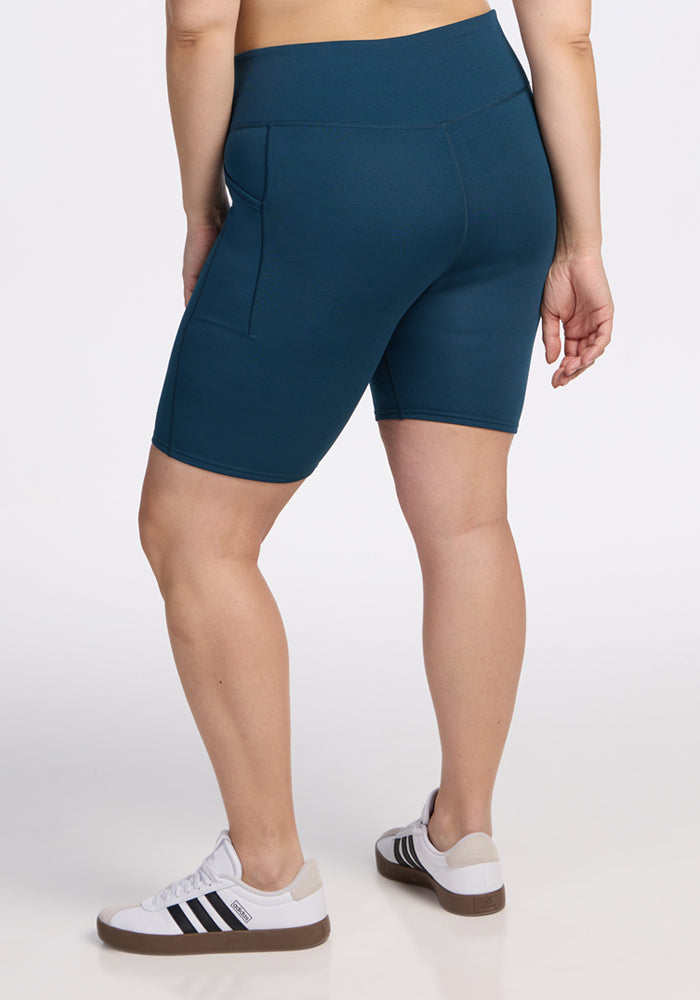 A person is wearing Woolx Merino Flex® Dani Bike Shorts in Majolica Blue with side pockets and white sneakers featuring black stripes, standing on a white background and facing away from the camera.