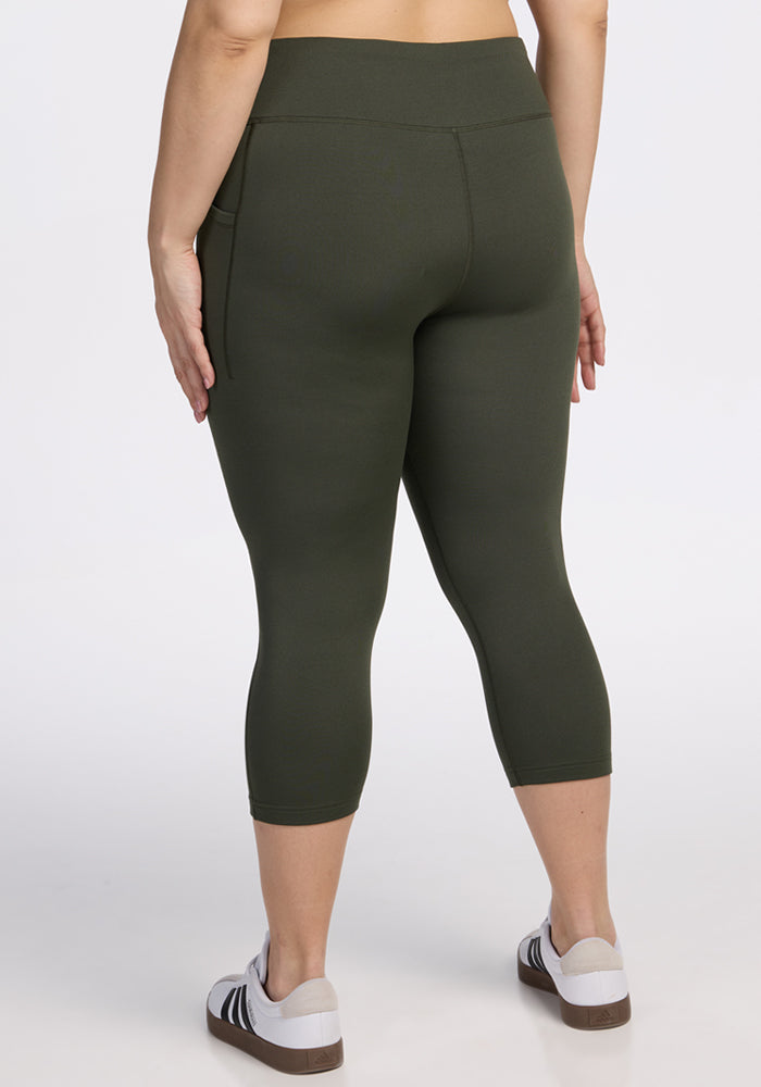 A person wearing Woolx Merino Flex® McKenna Capris in Everleaf and white sneakers with black stripes stands with their back to the camera against a plain white background.