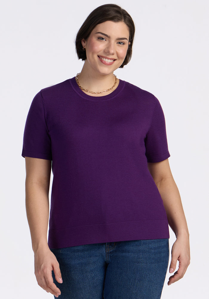 A woman with short brown hair smiles while wearing the Woolx Anna Crew Sweater in Plum Crush, paired with blue jeans and a gold chain necklace, standing against a plain white background. 