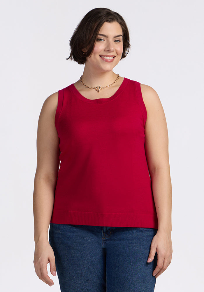 A woman with short brown hair wearing the Woolx Chessy Tank Top in Poppy Spark, blue jeans, and a necklace is smiling while standing against a plain white background. 