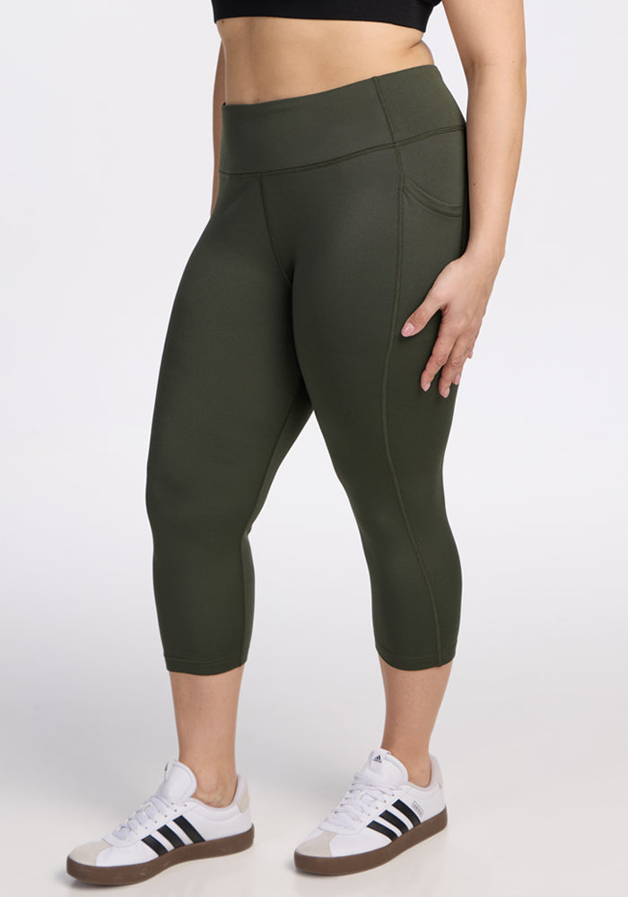 A person wearing Woolx Merino Flex® McKenna Capris in Everleaf and white Adidas sneakers with black stripes stands against a plain white background, showing only the lower torso and legs. 