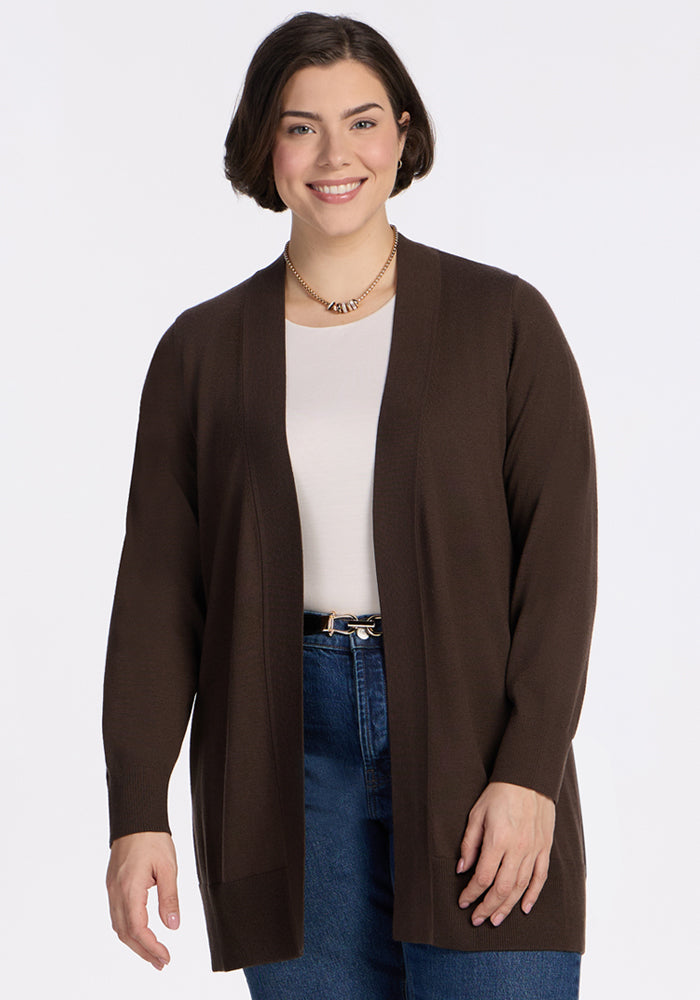 A woman with short dark hair smiles against a white background, wearing the Woolx Macie Sweater in Bracken over a white top and blue jeans, accessorized with a gold necklace. 