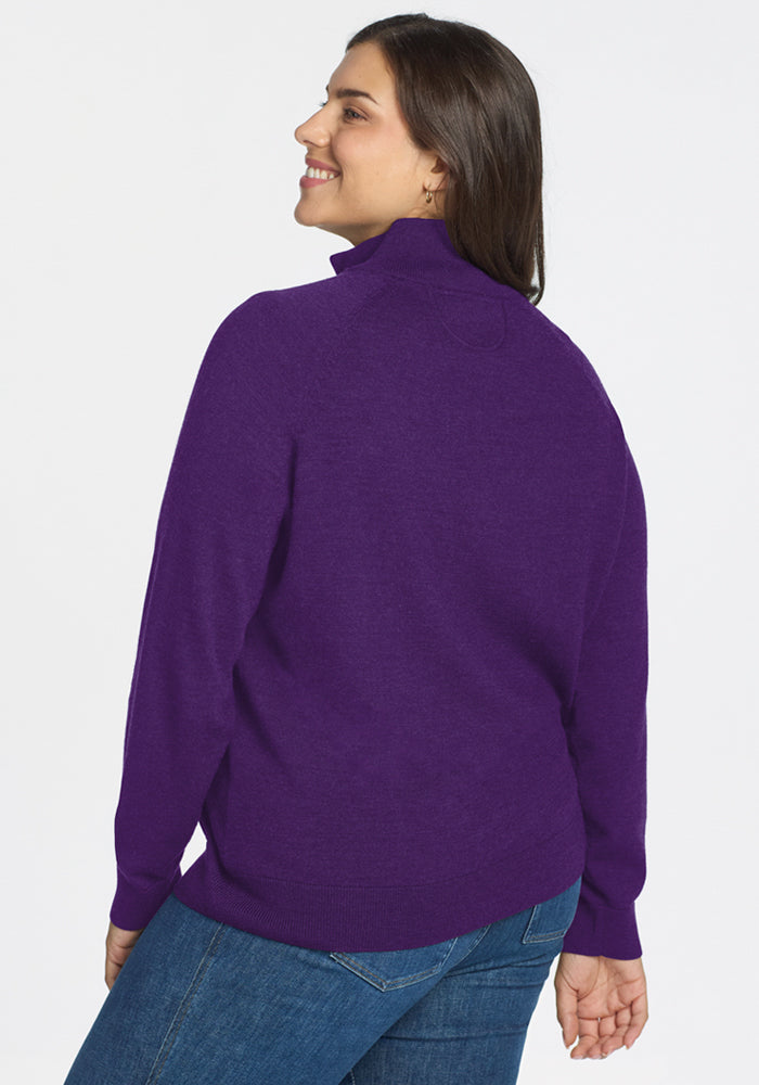 A woman with long brown hair smiles over her shoulder, wearing the Woolx Collins Half Zip Sweater in Plum Crush and blue jeans, standing against a plain white background.