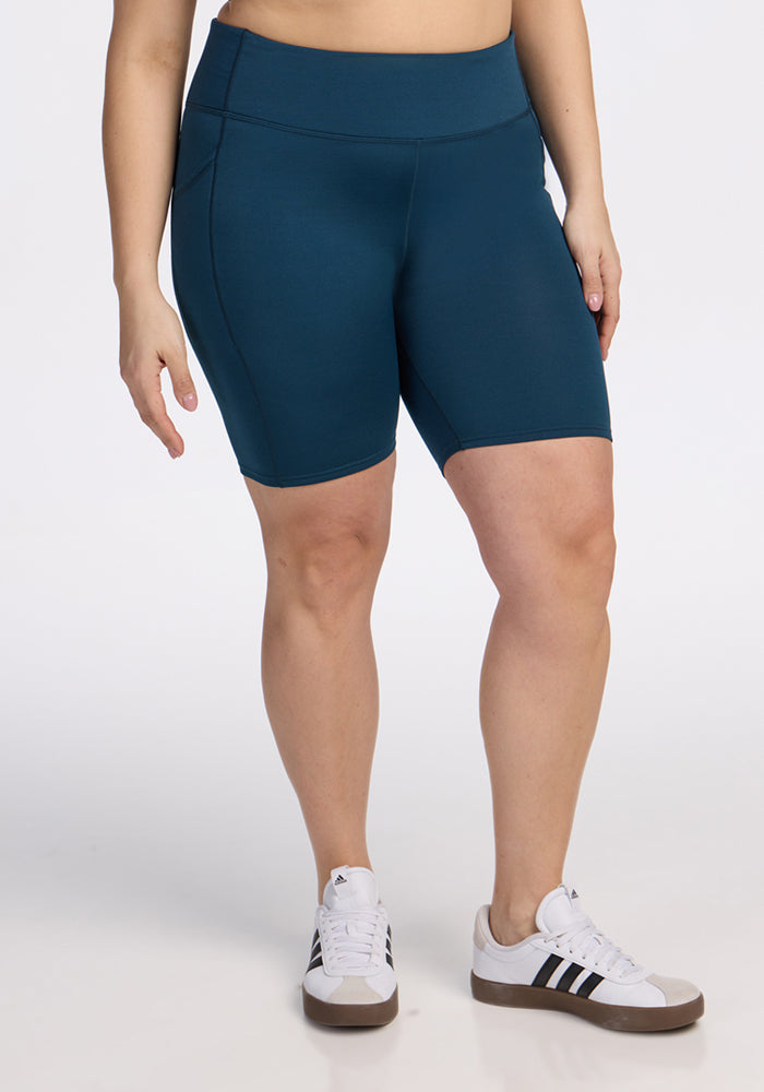 A person wearing Woolx Merino Flex® Dani Bike Shorts in Majolica Blue and white Adidas sneakers with black stripes stands against a plain white background. Their upper body is not visible. 