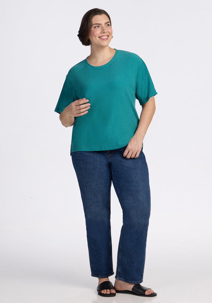 A smiling person wears the Woolx Delia Ribbed Short Sleeve in Surfside, blue jeans, and black sandals on a white backdrop.
