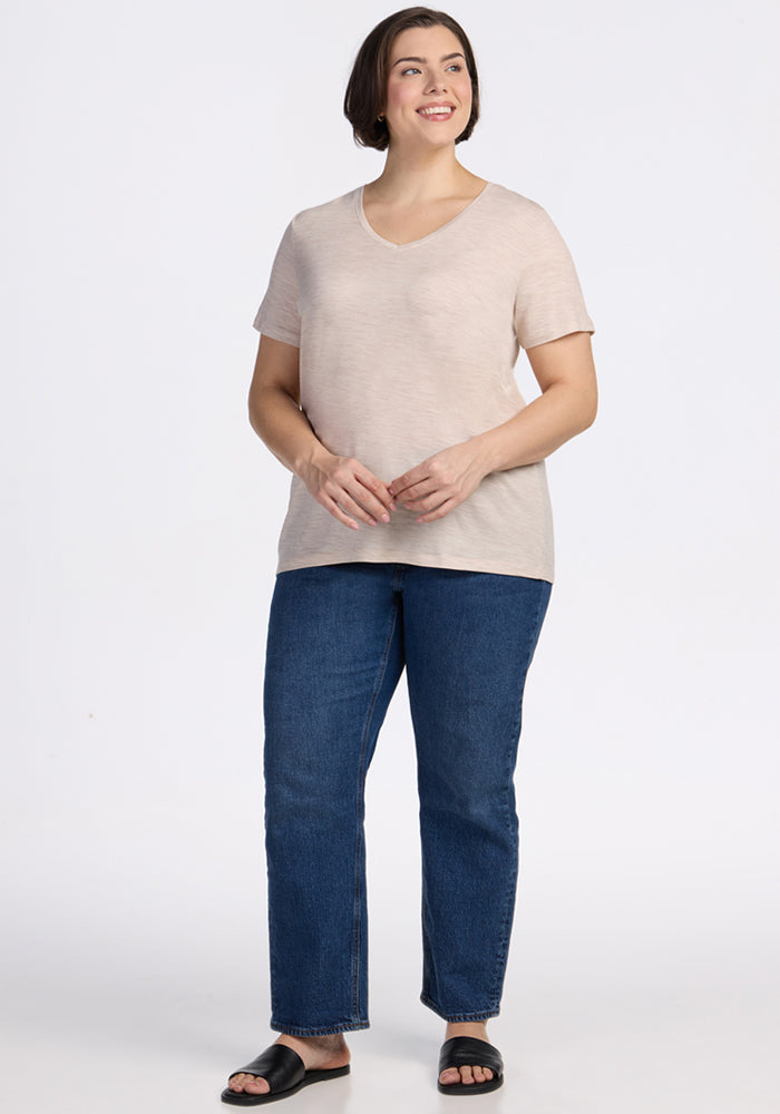 A woman with short brown hair smiles while standing against a white background, wearing the Woolx Junie V Neck in Cream Heather, blue jeans, and black sandals.