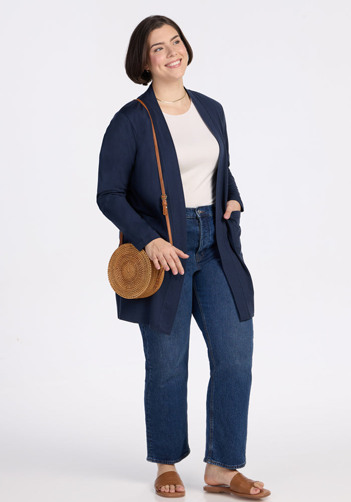 A woman with short dark hair wears the Woolx Paisley Cardigan in Deep Navy over a white shirt, blue jeans, and brown sandals. She carries a round straw crossbody bag and smiles against a plain white background.