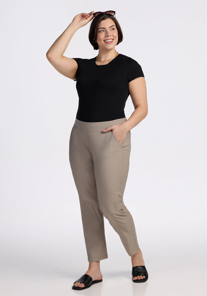 A woman with short dark hair wears a black t-shirt, Woolx Nicole Ankle Pants in Vintage Khaki, and black sandals, smiling against a white background as she holds sunglasses above her head with one hand and the other in her pocket.