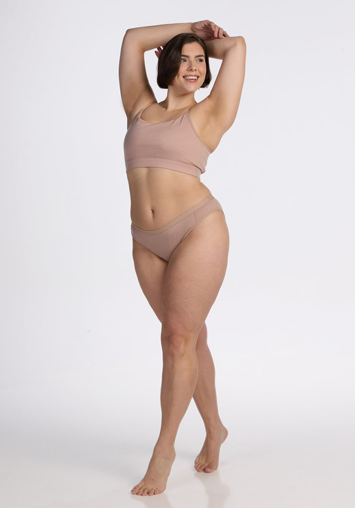 A woman with short brown hair smiles barefoot, posing with one arm raised in the Woolx Luna Bralette - Morning Latte, against a plain white background.