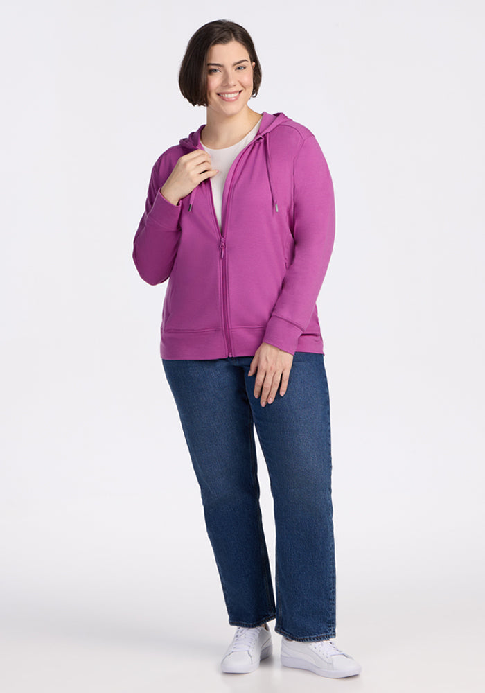 Smiling woman in a Woolx Ryann Hoodie • Final Sale - Purple Wine, blue jeans, and white sneakers against a white background.