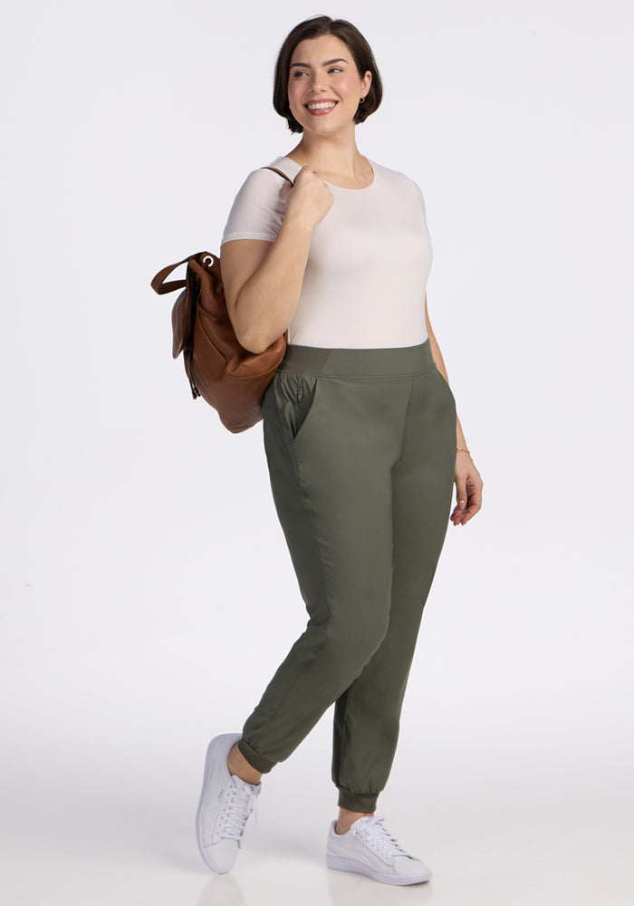 A woman smiles against a white background, wearing Woolx Palmer Joggers in Sage Olive, a light beige t-shirt, white sneakers, and a brown backpack over her shoulder with one hand in her pocket.