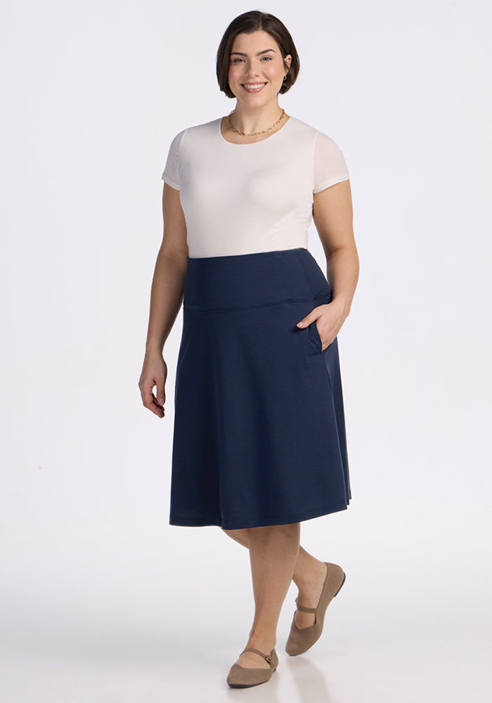 A woman with short brown hair wears a white short-sleeve top and the Woolx Marilyn Flowy Midi Skirt in Deep Navy. She stands facing forward, smiling in tan flats against a plain white background.
