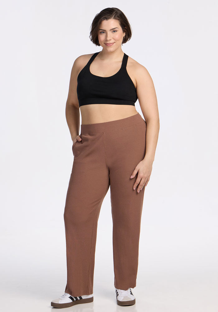 A woman with short brown hair wears the Woolx Luca Ribbed Pants in Sienna Kiss, a black sports bra, and white sneakers with black stripes. She stands confidently with one hand in her pocket against a plain white background.