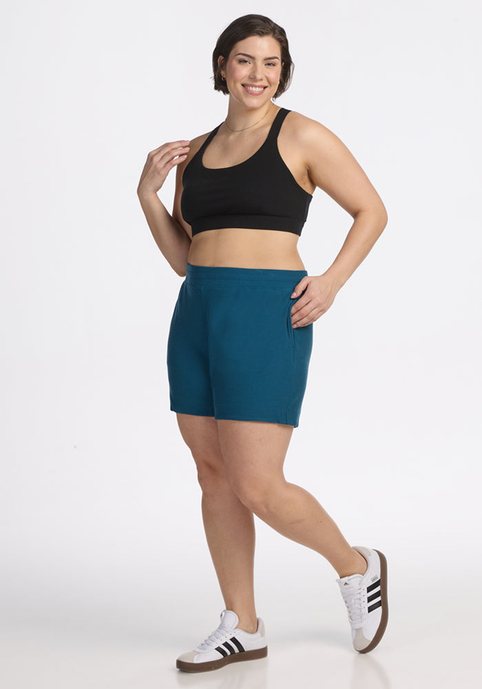 Woman smiling in Woolx Maisey Ribbed Shorts, Moroccan Blue, with a black sports bra and white sneakers on a white backdrop.