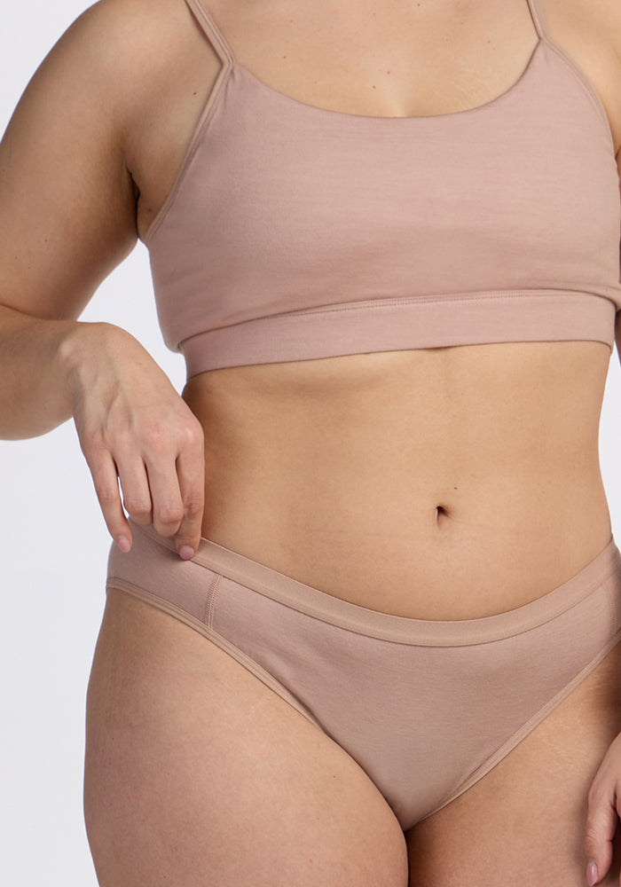 A person wears the Woolx Luna Bralette in Morning Latte with matching nude panties, standing against a plain background with one hand on their hip.