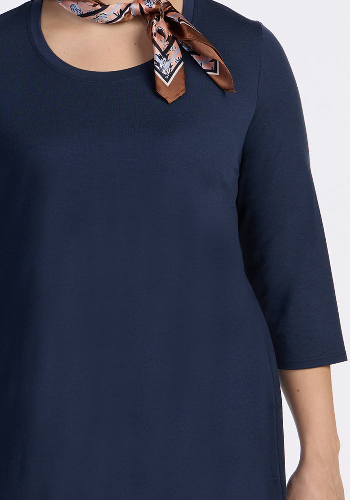 Someone wearing the Woolx Rissa A Line Dress in Deep Navy, styled with a brown patterned scarf at the neck—an ideal look for fall and winter layering. The background is plain and light-colored.