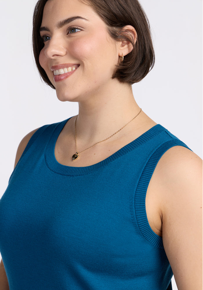 A woman with short brown hair smiles while wearing the Woolx Chessy Tank Top in Moroccan Blue, a small gold hoop earring, and a delicate gold pendant necklace against a plain light background.