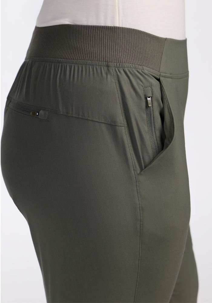 Close-up side view of a person wearing Woolx Palmer Joggers in Sage Olive, featuring an elastic waistband, zippered side pocket, and small back pocket. The lightweight joggers are paired with a light-colored top.