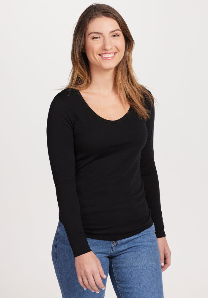 A person with long hair smiles while wearing the odor-resistant, merino wool Layla V Neck Top in black from Woolx, paired with blue jeans. The background is plain white. 