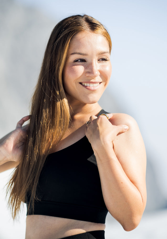 A woman with long light brown hair smiles outdoors in the sunlight, looking relaxed and happy, wearing the Woolx Livi Bra—a black, supportive one-shoulder style—with a blurred natural background behind her.