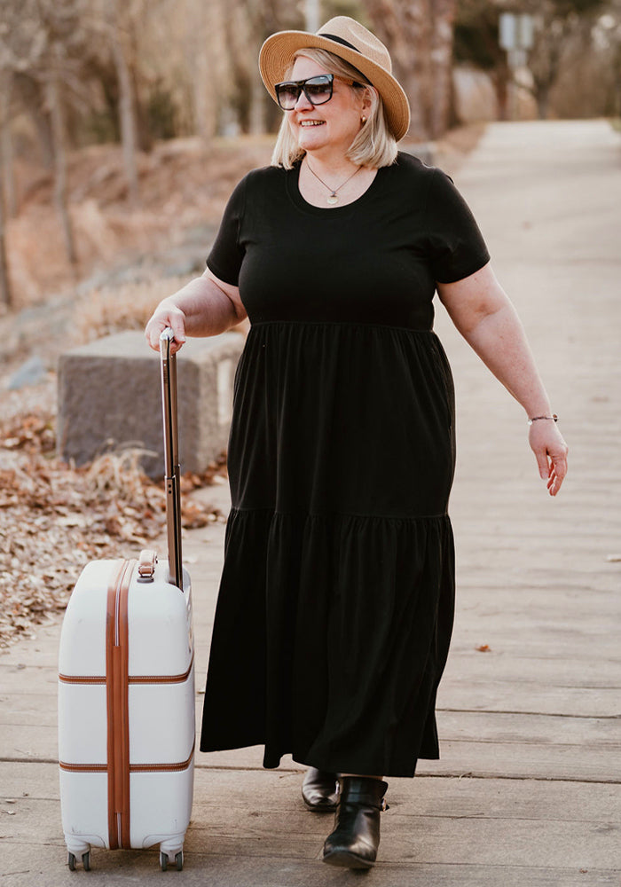 A smiling woman in a Woolx Lucia Dress - Black and hat walks outdoors with a white suitcase on a tree-lined path.