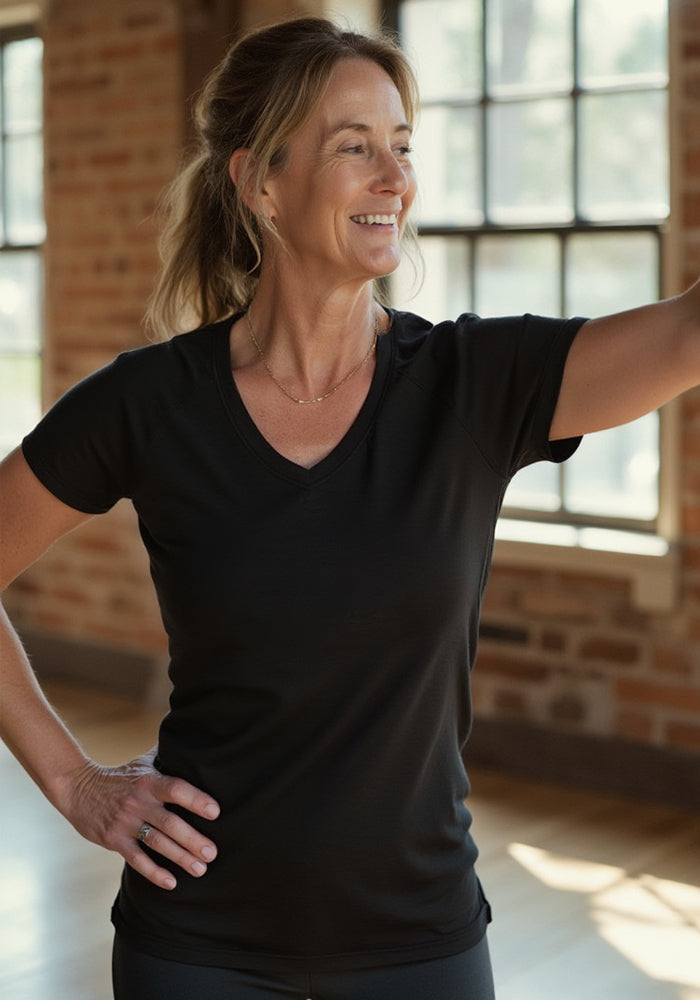 A smiling woman stands indoors by large windows, wearing the Woolx Mia V Neck in Black. Sunlight highlights her face and showcases the comfort of this moisture-wicking Merino wool t-shirt as she waves and poses with one hand on her hip.