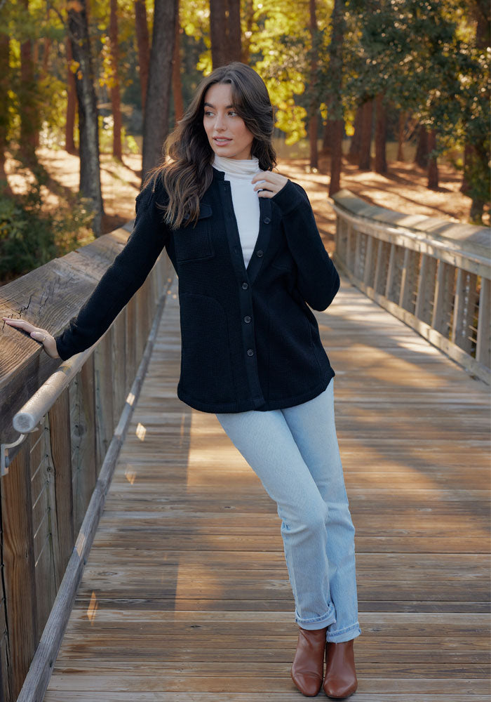 A woman with long brown hair stands on a wooden bridge in a forest wearing the Woolx Oaklynn Waffle Shacket in Biscotti over a white turtleneck, light blue jeans, and brown boots, gazing to the side with one hand on the railing.