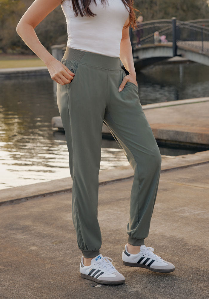 A person wearing a white sleeveless top, Woolx Palmer Joggers in Sage Olive, and white Adidas sneakers stands outdoors by a body of water and a bridge, with hands in their pockets.