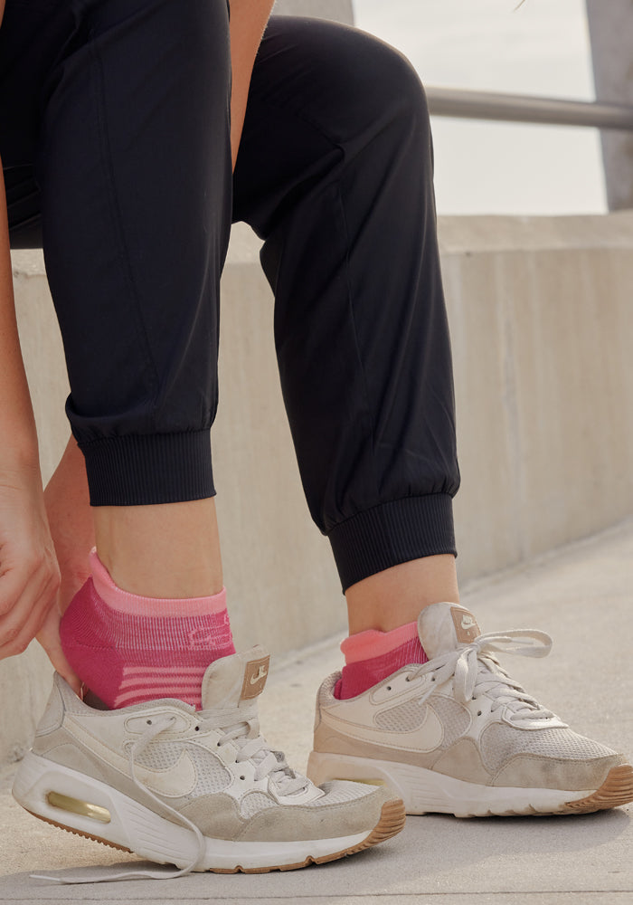 Wearing Woolx Palmer Joggers in black, a person adjusts bright pink socks above white Nike sneakers while sitting on a concrete bench outdoors.