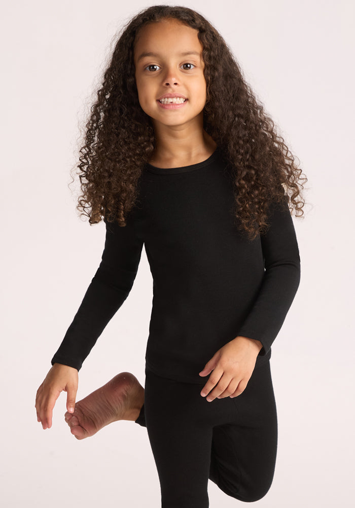 A young child with long curly hair wears the Woolx Kids Explorer Base Layer Top in black and matching pants, standing on one foot and smiling at the camera against a plain light background. 