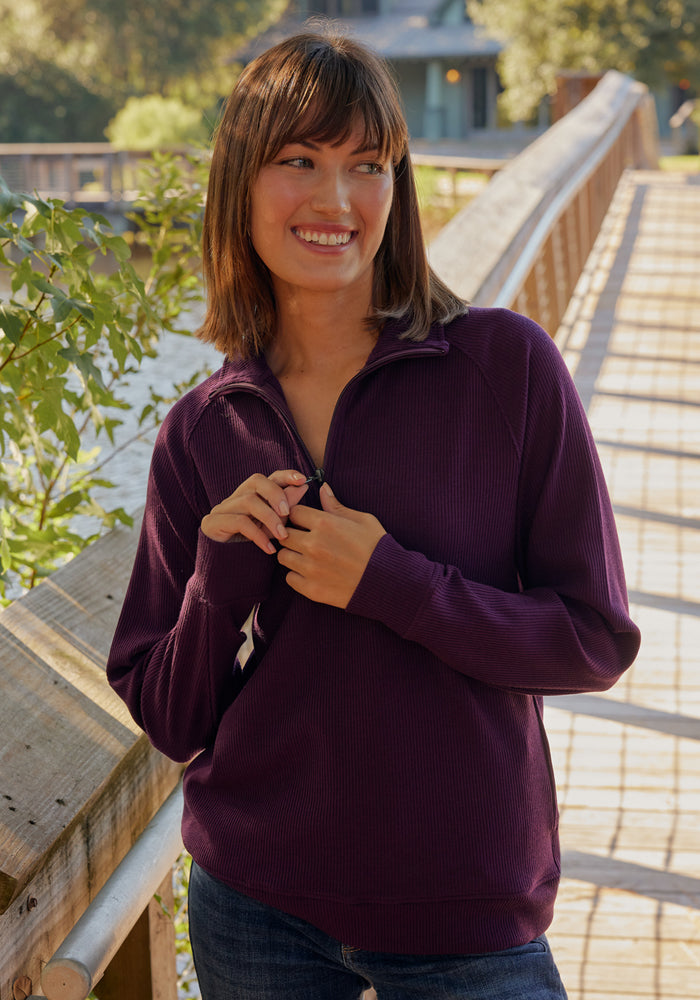 A woman with shoulder-length brown hair smiles on a wooden bridge outdoors, wearing the Woolx Quinn Ribbed 1/4 Zip in black and blue jeans, with greenery and sunlight in the background.
