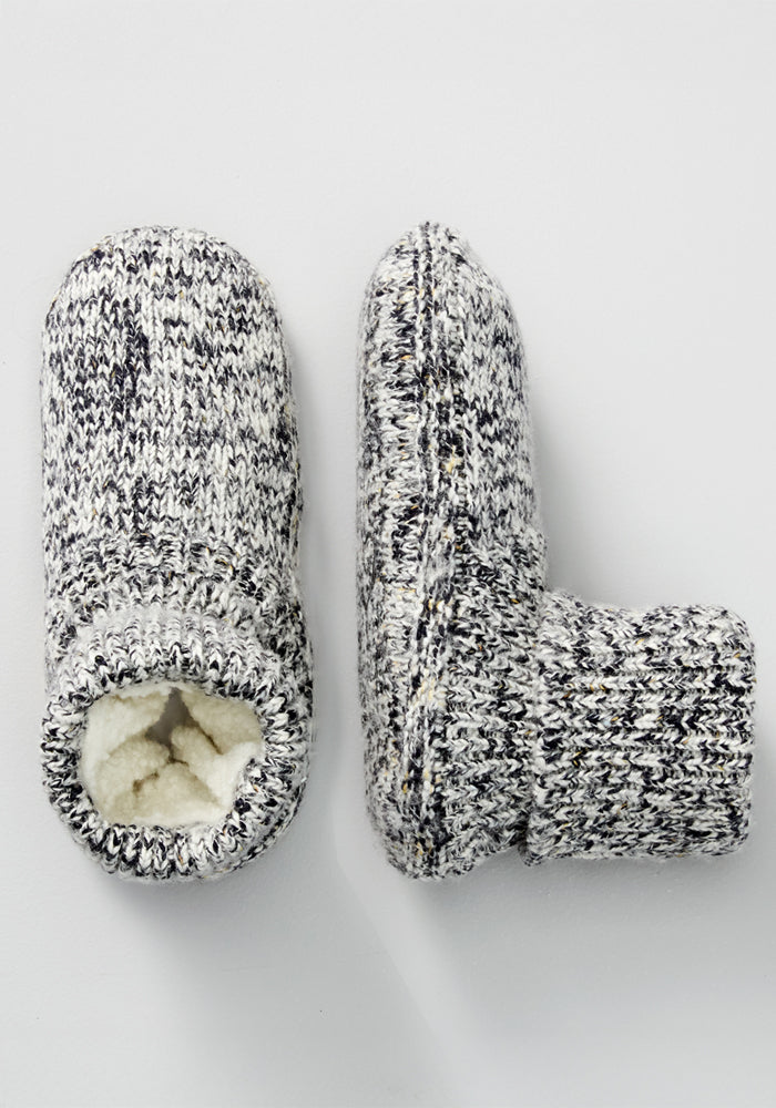 The Woolx Women's Gripper Slipper Booties in Speckled Stone feature cozy Merino wool, a soft white fleece lining, and a gray-and-white speckled design. One bootie stands upright while the other rests on its side to reveal the plush interior.