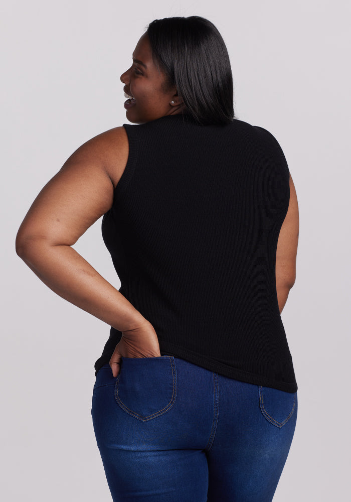 A person with long dark hair smiles over their shoulder, wearing a sleek black Raegan Ribbed Tank Top by Woolx and blue jeans, one hand in a pocket against a simple gray backdrop.