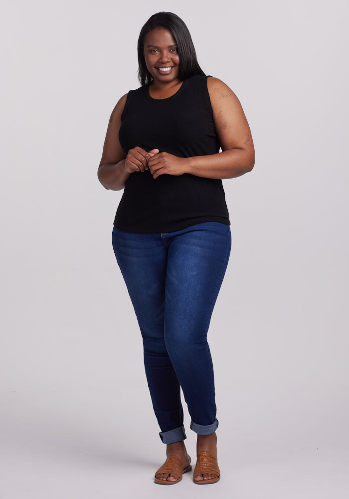 A person with long hair smiles confidently, wearing the Raegan Ribbed Tank Top by Woolx, blue jeans, and brown sandals. They stand against a plain gray background with hands gently clasped in front.