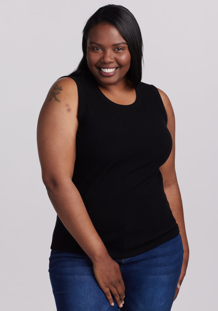 A person with long black hair smiles warmly, dressed in a sleek Woolx Raegan Ribbed Tank Top - Black and blue jeans. They have a tattoo on their left upper arm against a plain gray background. 