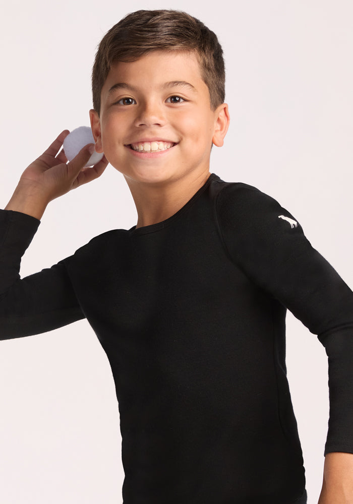 A smiling boy with short brown hair, wearing the Woolx Kids Explorer Base Layer Top in black, stands against a plain light background. He faces forward with his arm raised, preparing to throw a white ball.