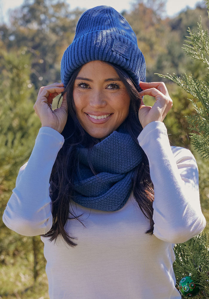 A woman smiles outdoors in a wooded area on a sunny day, wearing the Woolx Ria Chunky Beanie in Starry Night and a matching blue scarf with a white long-sleeve shirt.
