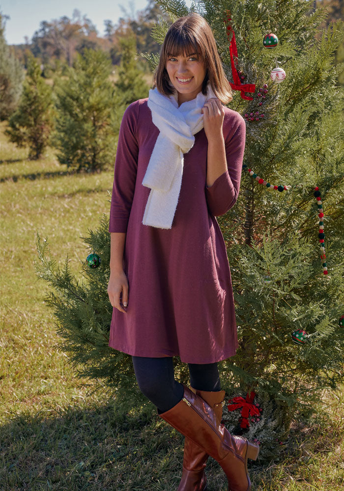 A woman in the Woolx Rissa A Line Dress in black, white scarf, black leggings, and brown boots stands smiling beside a decorated outdoor Christmas tree in a grassy field with evergreen trees behind her.