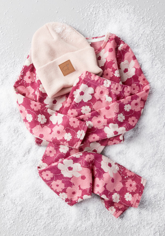 The Woolx Kids Explorer Base Layer Bottoms in Rosie Posie, paired with a pink knit beanie, are arranged on snow—perfect cozy wear for kids to stay warm and stylish during chilly outdoor adventures.